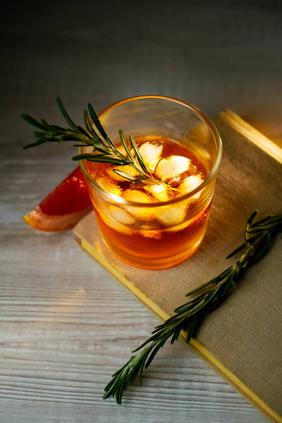 Whiskey sour cocktail with rum ice grapefruit and rosemary in shot glass on book. Alcohol cocktail in light beam - 写真・画像