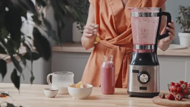 PAN mid-section of young woman putting straw into glass bottle with freshly made smoothie in cozy kitchen. Ingredientes frescos e leite em jarro estão no balcão - Filmagem, Vídeo