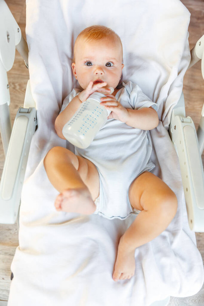 Cute little newborn girl drinking milk from bottle and looking at camera on white background. Infant baby sucking eating milk nutrition lying down on feeding chair at home. Motherhood happy child - Foto, Bild