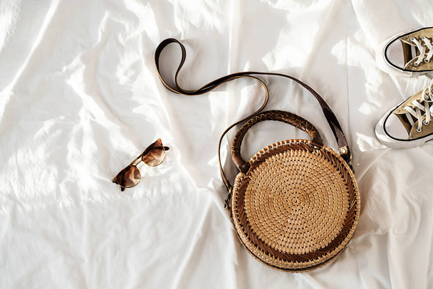 Trendy round rattan woven bag, sunglass and sneakers in sunlight with shadows, top view. Summer or spring fashion concept. - Photo, Image