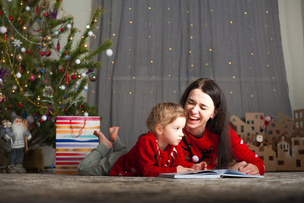 Nuori onnellinen äiti ja tytär punaisissa villapaidoissa lukevat kirjoja kotona. Joulu tunnelma, DIY sisustus pahvista... - Valokuva, kuva