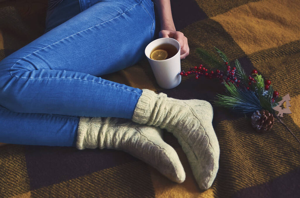 A girl in blue jeans and woolen knitted socks holds a glass of tea in her hands, close-up. Lifestyle concept. - Φωτογραφία, εικόνα