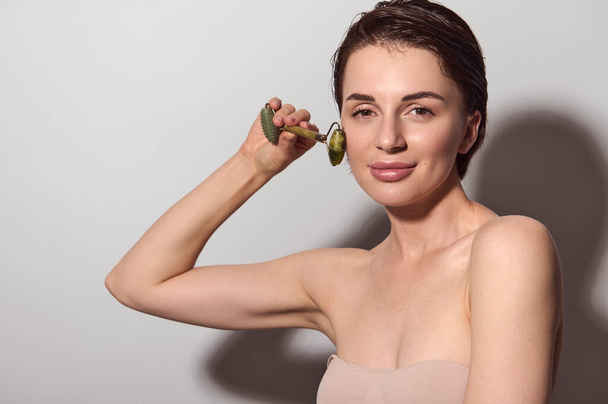 Skin care, anti-aging, facial lifting massage and wellness spa concept. Close-up portrait of a natural beauty Caucasian woman massaging her face with jade roller stone massager. - Foto, Imagem