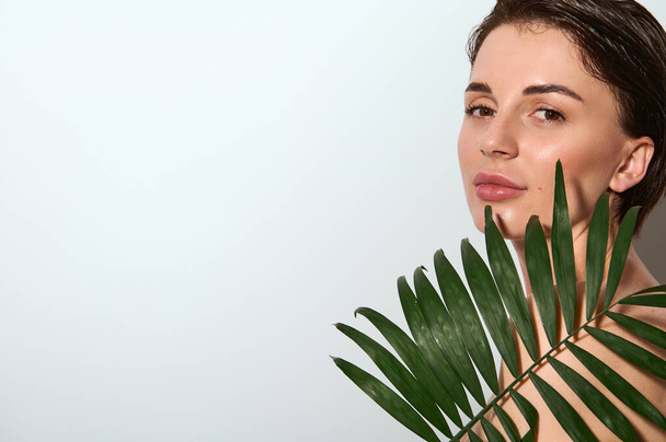 Egzotik palmiye yaprağı olan, beyaz arka planda fotokopi aletiyle poz veren muhteşem, çekici, çıplak bir kadının yatay yakın çekimi. Cilt bakımı için doğal organik kozmetik kullanımı kavramı - Fotoğraf, Görsel