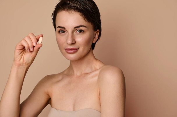 Close-up. Attractive European naked natural beauty woman with healthy shiny skin, holding a pill with dietary supplement, complex of vitamin and minerals in dragee. Medicine, pharmacy, cosmetology - Photo, image