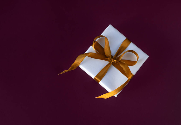 Caja de regalo envuelta en papel blanco con un lazo dorado sobre fondo violeta festivo - Foto, imagen