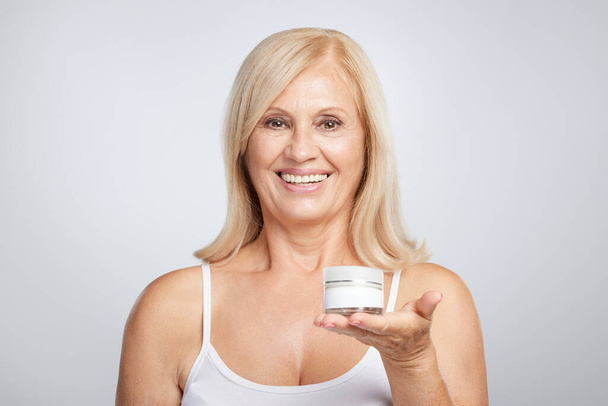 Una mujer rubia mayor feliz sosteniendo crema anti-edad en la mano y mirando a la cámara. - Foto, imagen