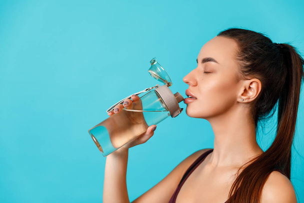 close-up portrait of a beautiful young brunette woman in a sportswear drinks water from a bottle on a blue background. space for text - Photo, Image