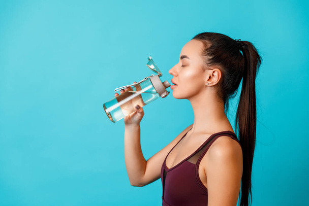 retrato de una hermosa joven morena en ropa deportiva bebe agua de una botella sobre un fondo azul. espacio para texto - Foto, Imagen