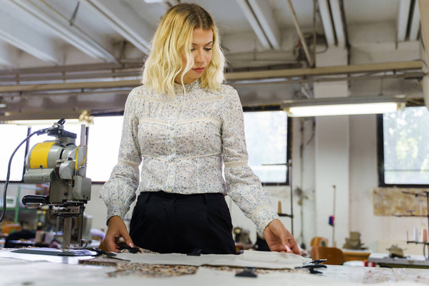 若い大人ブロンドの白人女性がテーラーショップで素材に新しいデザインのスケッチドレスを作る-テーブルによって働く女性起業家-中小企業の男女共同参画の概念 - 写真・画像