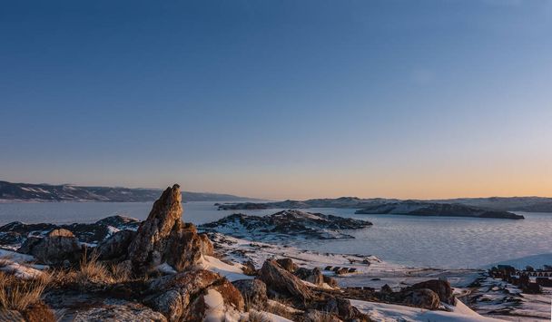 Kora téli reggel Szibériában. Az ég narancssárgával van kiemelve. Egy befagyott tó jegén süt a nap. Kempingezni egy havas parton. Az előtérben festői gránitsziklák, száraz fű található. Bajkál.  - Fotó, kép