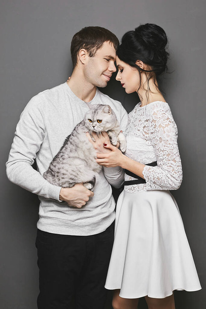 Sweet couple of lovers, a young gorgeous woman in dress and handsome guy posing with their cute cat on the grey background - Foto, Bild