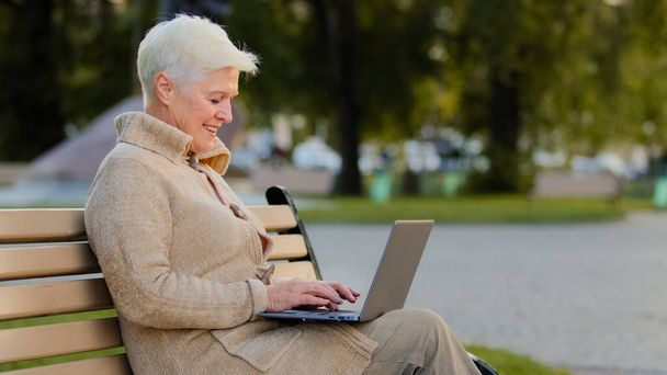 Aged woman sitting on bench in city park using computer surfing internet, smiling senior female of retirement age working at laptop reading news online, elderly lady texting message at pc outdoor - Фото, изображение