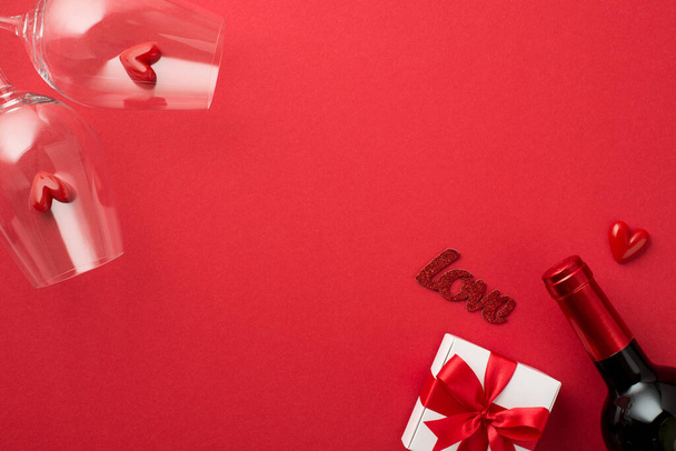 Widok z góry zdjęcie Walentynki dekoracje biały prezent pudełko z czerwonym łukiem małe serca w dwóch kieliszków z napisem miłość i wino butelka na izolowanym czerwonym tle z pustym miejscem - Zdjęcie, obraz