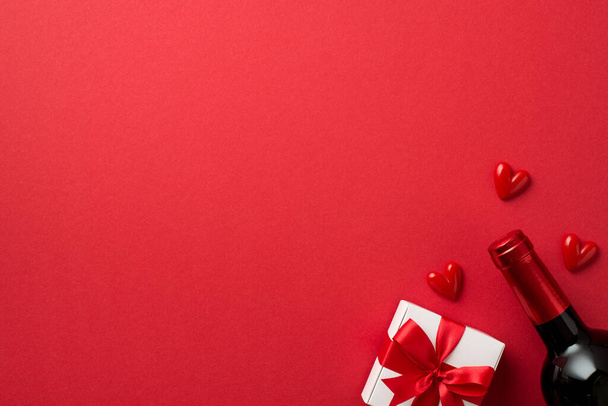 Вид зверху фото прикрас дня Валентина біла подарункова коробка з червоним бантом маленькі серця та пляшка вина на ізольованому червоному тлі з порожнім простором
 - Фото, зображення