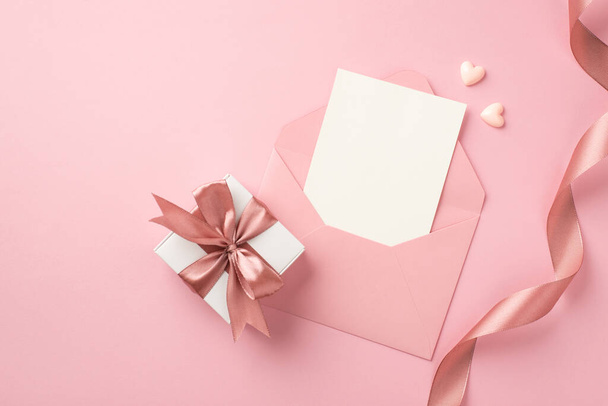 Top view photo of white giftbox with pink bow small hearts curly silk ribbon and open pink envelope with paper card on isolated pastel pink background with blank space - Foto, Bild