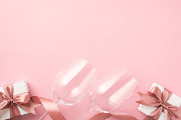 Photo de vue du dessus des décorations de la Saint-Valentin deux verres à vin ruban de satin rose et boîtes-cadeaux blanches avec des arcs sur fond rose pastel isolé avec copyspace - Photo, image