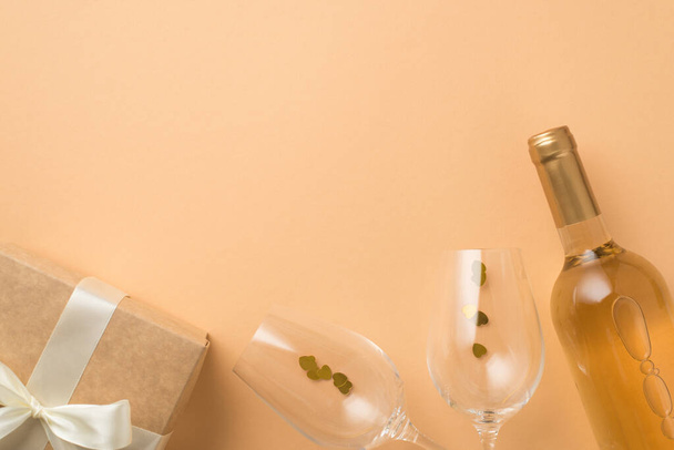 Top view φωτογραφία του Αγίου Βαλεντίνου διακόσμηση μπουκάλι λευκό κρασί δύο ποτήρια κρασί με κονφετί σχήμα καρδιάς και craft χάρτινο κουτί δώρου με λευκό τόξο σε απομονωμένο παστέλ μπεζ φόντο με copyspace - Φωτογραφία, εικόνα