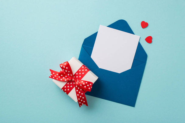 Bovenaanzicht foto van open blauwe envelop met papieren velletje klein rood hartje en wit geschenkdoosje met rood lint strikje op geïsoleerde pastelblauwe achtergrond met lege ruimte - Foto, afbeelding