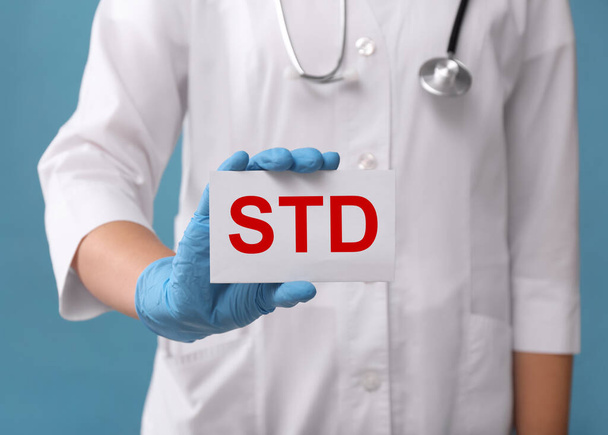 Doctor holding sheet of paper with abbreviation STD on light blue background, closeup - Zdjęcie, obraz