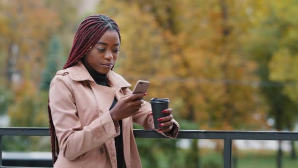 Ελκυστική νεαρή Αφρο-Αμερικανίδα φοιτήτρια πίνοντας καφέ χαλαρό υπαίθριο μόνο την ημέρα του φθινοπώρου. Μακριά μαλλιά χιλιετή γυναίκα κρατώντας το κινητό τηλέφωνο κοιτάζοντας οθόνη smartphone κουβεντιάζοντας στα μέσα κοινωνικής δικτύωσης - Πλάνα, βίντεο