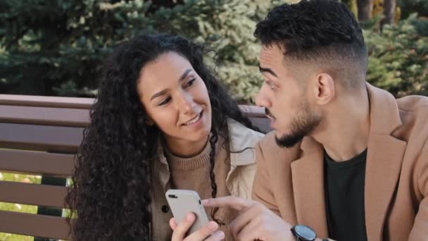 Hispánský pár sedět na lavičce v podzimním parku mladý chlap držet telefon muž a dívka mluvit pohled na obrazovce zařízení překvapený rodina usmívá pomocí aplikace, aby on-line objednávky muž aktivně komunikovat s přítelkyní - Záběry, video