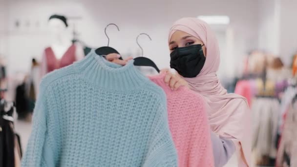 Musulman jeune femme en masque de protection médicale pendant la quarantaine pandémique fille islamique en hijab vendeur dans le magasin de vêtements propose des pulls en vente discount acheteur féminin ne peut pas choisir entre deux couleurs - Séquence, vidéo