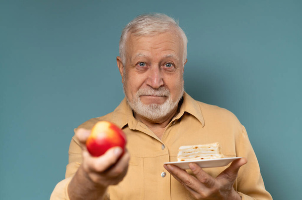 A man with gray hair holding a cake and an apple, to choose from, healthy food concept, on a blue background - Photo, Image