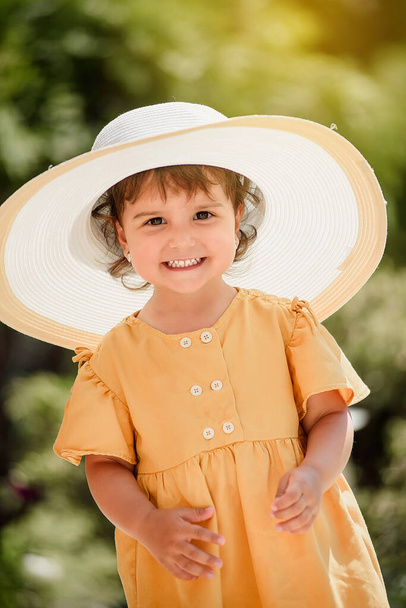 Happy smiling little girl kid in yellow dress is wearing an oversize white hat and looking to the camera - Foto, immagini