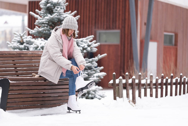 A girl laces up her skates while sitting on a park bench. A young woman came to skate at the rink. - Foto, afbeelding