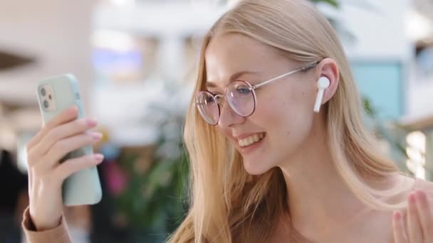 Young millennial girl make video call in mall wave hand say hello friend smiling female in wireless headset happy caucasian woman having fun talk use phone virtual meeting remote communication concept - Felvétel, videó