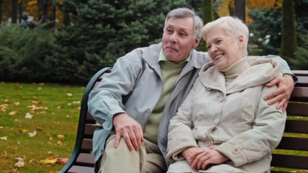 Šťastný dospělý ženatý pár usměvavý sedí na lavičce v podzimním parku starší manžel objímání milovaná manželka rodina těšit konverzace venku radostné důchodci objímat pozitivní zralé manželky sdílet zprávy - Záběry, video