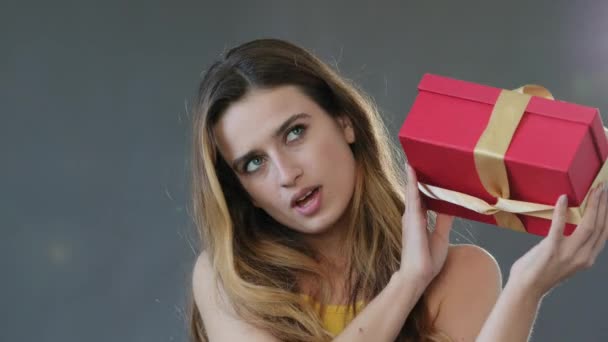 Любопытная молодая женщина смешной кавказка женщина модель день рождения девушка, изолированная на сером фоне студии встряхивает красный подарочная коробка с сюрпризом слушать подарок внутри, поздравления праздничной концепции - Кадры, видео