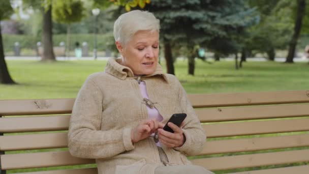 Mujer madura de pelo gris muy alegre sentada al aire libre, utilizando aplicaciones de teléfonos inteligentes de navegación, mirando a la pantalla, mujer mayor charlando con familiares en línea, compras, divertirse con el dispositivo móvil - Imágenes, Vídeo