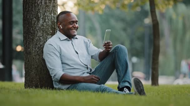 Elderly man entrepreneur sitting on grass in city park businessman holding smartphone looking at screen during video chat emotionally talks shares good tidings great news with partner  - Filmagem, Vídeo