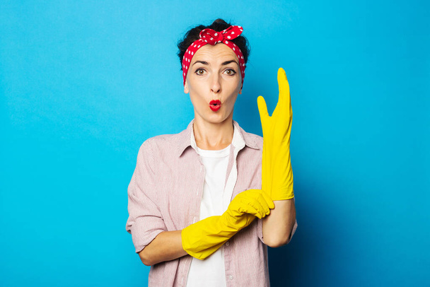 Surprised young woman in shirt with cleaning gloves on blue background - Фото, зображення