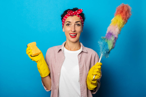 Smiling young woman in gloves holding a sponge and dust brush on blue background - Photo, image