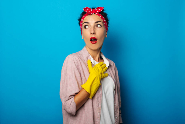 Sprzątanie. Zaskoczona młoda kobieta z bandażem na włosach w rękawiczkach czyszczących na niebieskim tle - Zdjęcie, obraz