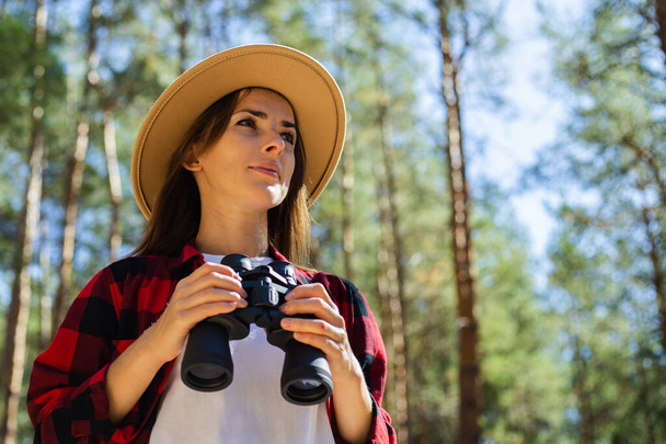 帽子の女性と森の中で双眼鏡を持っている赤い再生シャツ. - 写真・画像
