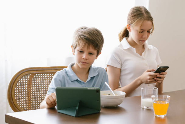 Bratr a sestra sedí během snídaně v kuchyni. Chlapec se dívá na digitální tablet a dospívající dívku kontrolující sociální média držící smartphone. Nadměrné používání zařízení, závislost gadgets koncept závislosti. - Fotografie, Obrázek