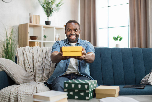 African man in denim shirt holding gift box on couch - Фото, изображение