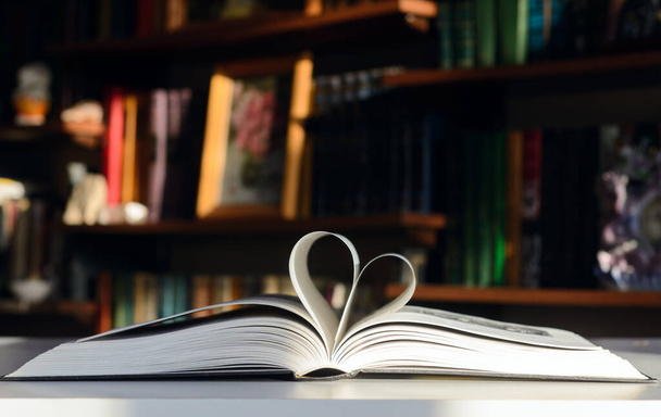 Background - bookshelves lit by the sun. An open book is on the table. Pages are folded in the shape of a heart.  Concept - Valentine's Day - Photo, Image