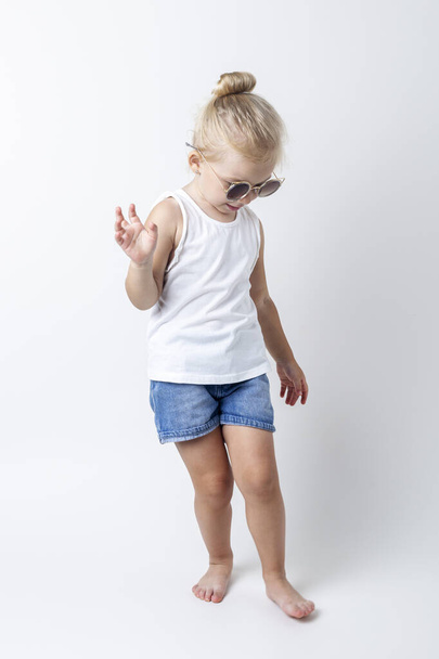 Szőke gyermek napszemüvegben pózol egy könnyű háttér a stúdióban. - Fotó, kép