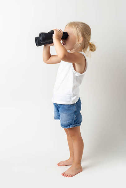 Blond dítě dívá dalekohledem na světlém pozadí ve studiu.  - Fotografie, Obrázek