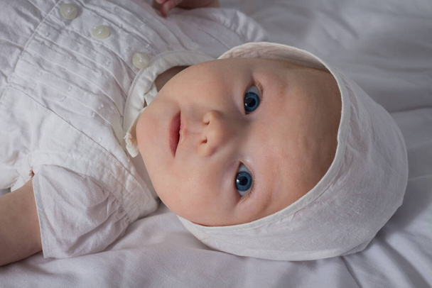 Κοριτσάκι με μπλε μάτια σε λευκό φόρεμα και καπέλο σε λευκή κουβέρτα. - Φωτογραφία, εικόνα