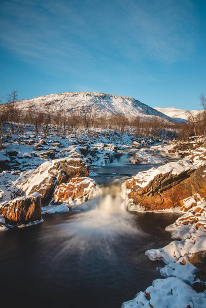 Zimowa bajka w postaci wodospadu Storforsen w Silsand, Senja, Norwegia. Zachód słońca oświetla dziki wodospad. W tle śnieżne wzgórza norweskiego krajobrazu. - Zdjęcie, obraz