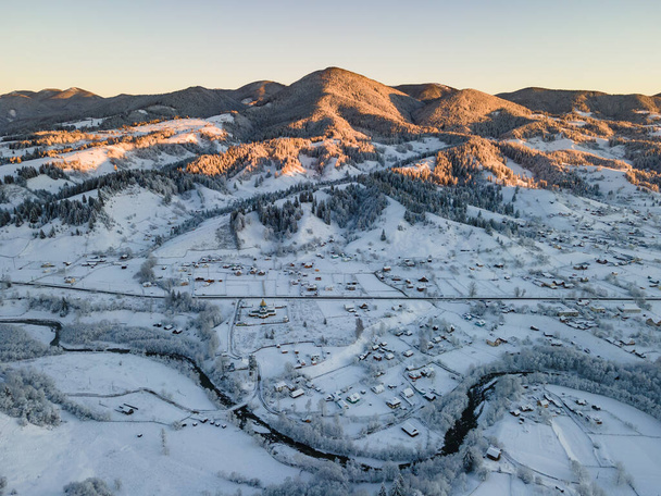 Aerial winter landscape with small village houses between snow covered forest in snow mountains - Fotó, kép