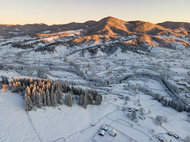 Luchtfoto winterlandschap met kleine dorpshuisjes tussen besneeuwd bos in koude bergen  - Foto, afbeelding