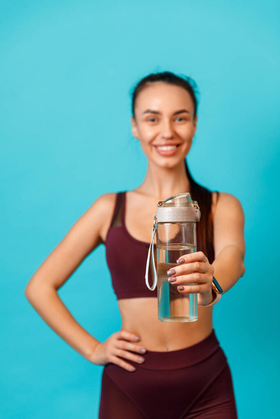 jonge brunette vrouw in sportkleding biedt u een fles met water op een blauwe achtergrond. fles in focus. - Foto, afbeelding