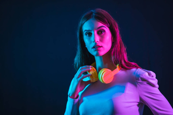 Young caucasian girl with yellow headphones on a black background, illuminated in a disco with neon lights - Foto, immagini